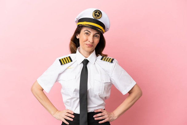 Avion pilote d'âge moyen femme isolée sur fond rose posant avec les bras à la hanche et souriant - Photo, image