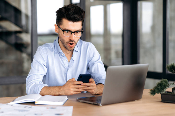 Pleasantly shocked caucasian man with, manager, broker or freelancer, sitting in the office, using mobile phone, surprised by news or message, looking at screen in amazement, tearing his mouth - Photo, Image
