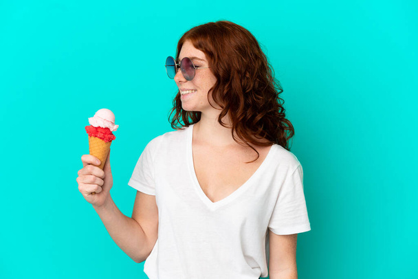 Teenager reddish woman with a cornet ice cream isolated on blue background looking side - Фото, зображення