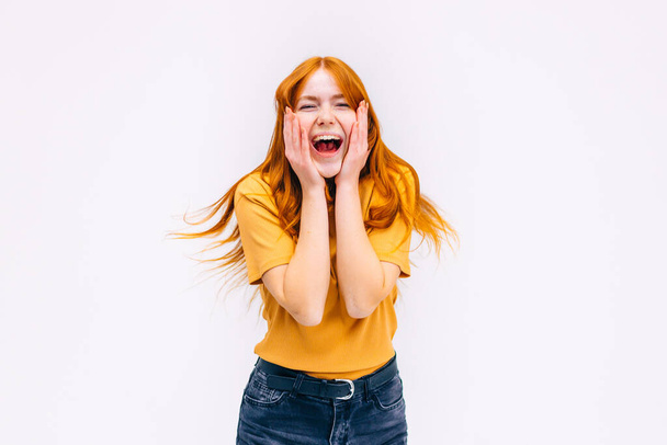Smiling overjoyed young ginger girl in yellow clothes jumping laughing showing winner gesture saying yes winning lottery extremely happy joyful isolated on white background in studio. Real emotions - Zdjęcie, obraz