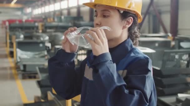 Naklápěcí pomalý portrét mladé ženské inženýrky v kombinéze a tvrdém klobouku, nasazující bezpečnostní brýle a pózující pro kameru stojící v továrně - Záběry, video