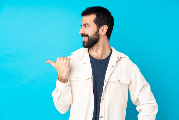Young handsome man with white corduroy jacket over isolated blue background pointing to the side to present a product - Photo, Image
