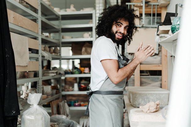  A curly-haired man crumples pottery clay with his hands in a hobby studio in an apron and a white t-shirt - Fotoğraf, Görsel