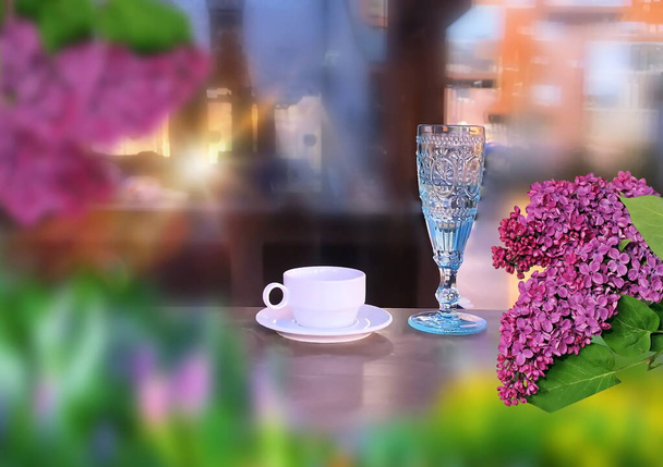  street food  lilac flowers white  cup of coffee and blue glass of wine on wooden table at street cafe sunlight reflection on windows city urban life stile - Photo, Image