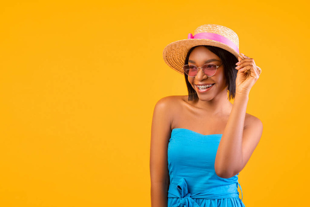 Vibras de verano. Retrato de la alegre joven negra en vestido, sombrero de paja y gafas de sol mirando el espacio en blanco - Foto, imagen