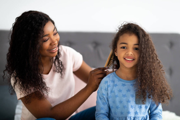 Beautiful black woman brushing her daighters hair on bed indoors - Foto, immagini
