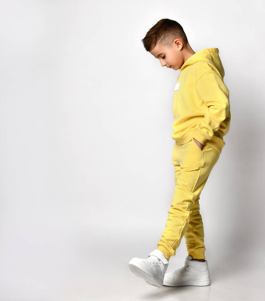 Profile view of a little brunette boy in yellow sportswear and white sneakers. - Foto, imagen