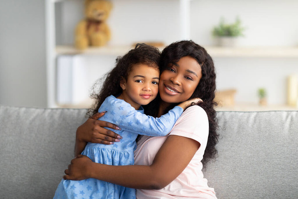 Kochająca czarna dziewczyna w ładnej sukience przytulająca matkę na kanapie w salonie. Koncepcja silnych relacji rodzinnych - Zdjęcie, obraz