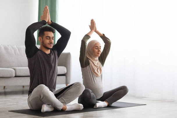 妊娠中のイスラム教徒のカップルは、自宅でヨガを練習,頭の上に手で座って - 写真・画像