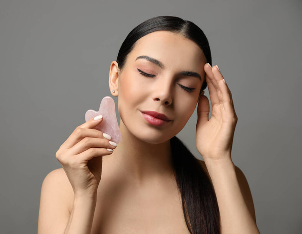 Beautiful young woman doing facial massage with gua sha tool on grey background - Fotoğraf, Görsel