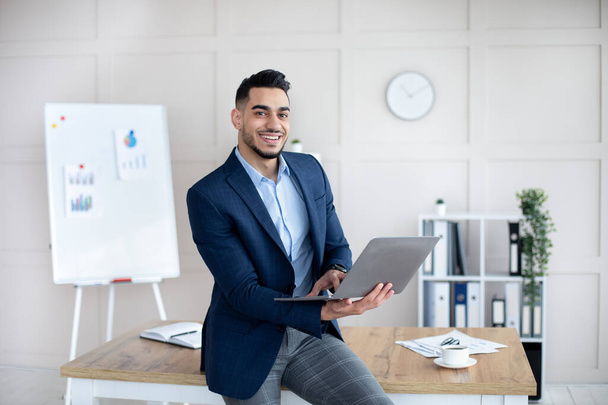 Lächelnder arabischer Geschäftsmann im formellen Anzug mit Laptop im Büro - Foto, Bild