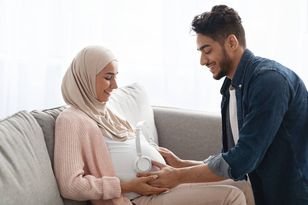 Milující arabský manžel umístění bezdrátových sluchátek na těhotné manželky břicho - Fotografie, Obrázek