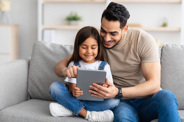 Smiling dad and daughter sitting on couch, using digital tablet - Fotoğraf, Görsel