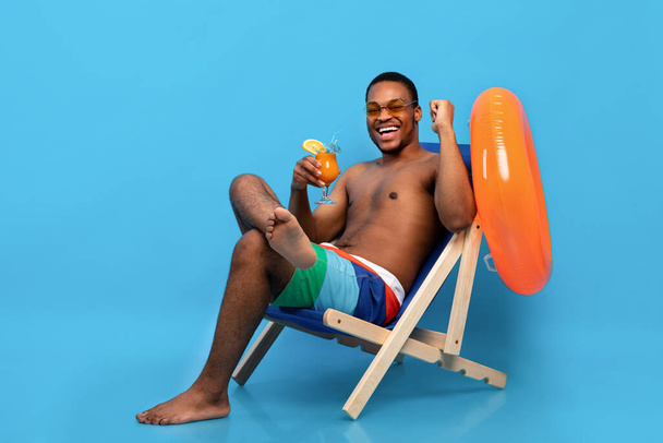 Volle Länge fröhlicher schwarzer Kerl mit tropischem Cocktail, der im Liegestuhl chillt und JA-Geste vor blauem Hintergrund macht - Foto, Bild