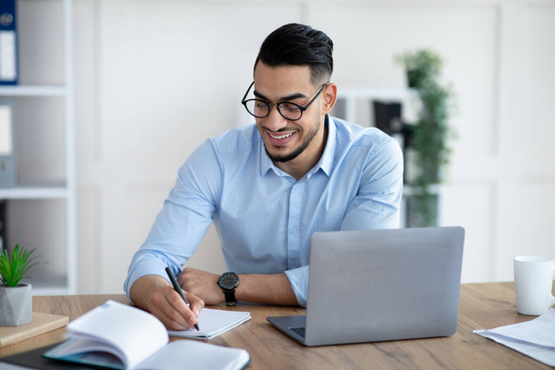 Arabische mannelijke ondernemer schrijft informatie op tijdens online zakelijke bijeenkomst op modern kantoor - Foto, afbeelding