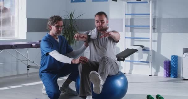 Therapeut van middelbare leeftijd helpt patiënt om evenwicht op fit bal - Video