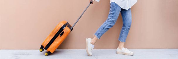Young Asian girl happy while towing her bright orange suitcase with right hand and left hand fist bumping in the air on concrete floor and desert sand colored wall in her background - Photo, Image