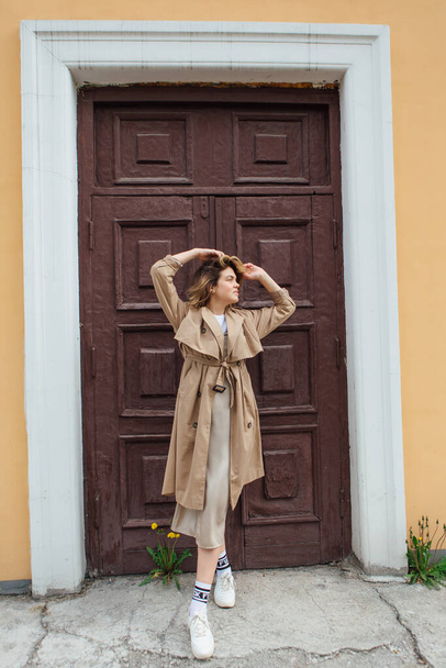 Young millennial woman with wild hair dressed in an autumn coat posing near the door of an old building. - 写真・画像