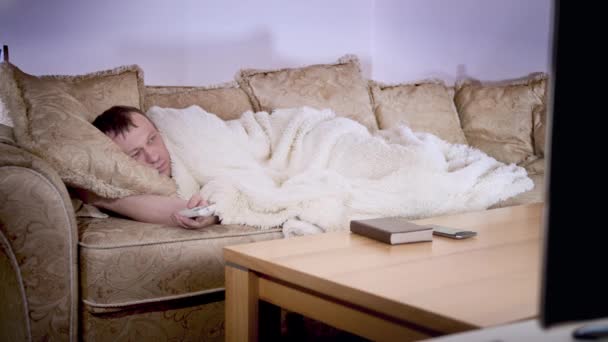 Un hombre yace cubierto con una manta en un sofá, interruptores de canales de televisión, iluminación nocturna - Metraje, vídeo