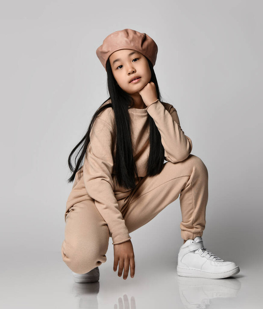 Active asian brunette kid girl in leather hat beret, cotton pants, hoodie, sweater and sneakers sits squatted - Фото, изображение