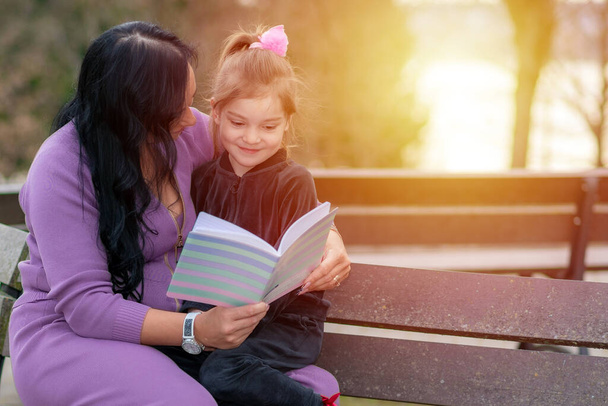 Mooi klein meisje dat een boek leest met haar moeder en glimlacht. Zomer wazig park in de achtergrond. vrouw het lezen van een boek aan haar dochter genieten van een warme avond in de lokale zonsondergang park - Foto, afbeelding