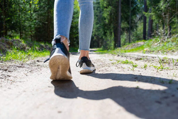 Achteraanzicht foto van vrouwelijke toeristische benen met sneakers wandelen in een bos weg.Zomer dag zon. - Foto, afbeelding