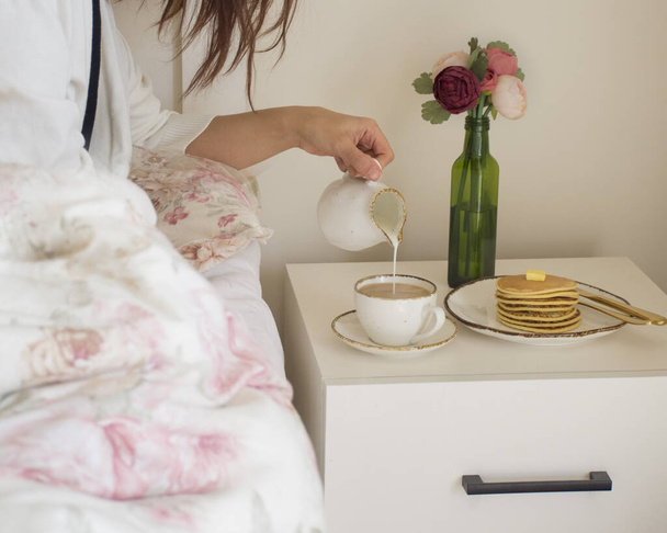 The picture shows a female hand pouring milk into her coffee. There is a stack of pancakes on the nightstand. - Foto, afbeelding