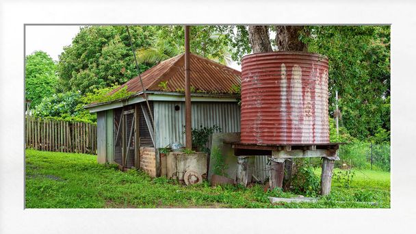 Oude schuur en watertank in een buitentuin na regenval wanneer het gras en gebladerte weelderig en groen zijn, met wit matframe - Foto, afbeelding