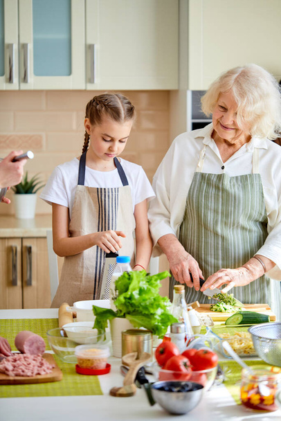 Happy adult senior lady teaching little teen girl how to cook healthy food - Photo, Image