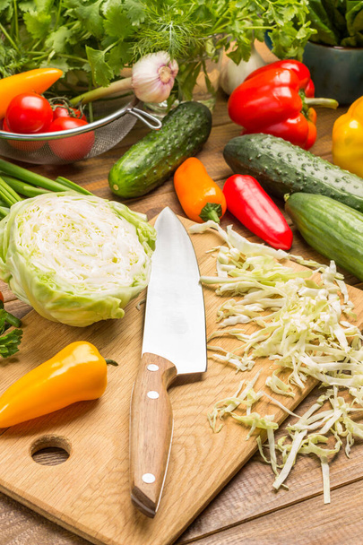 Sliced cabbage, kitchen knife on cutting board. Peppers, cucumbers, garlic and tomatoes and dill, spinach leaves and cilantro on table. Wooden background - Photo, image