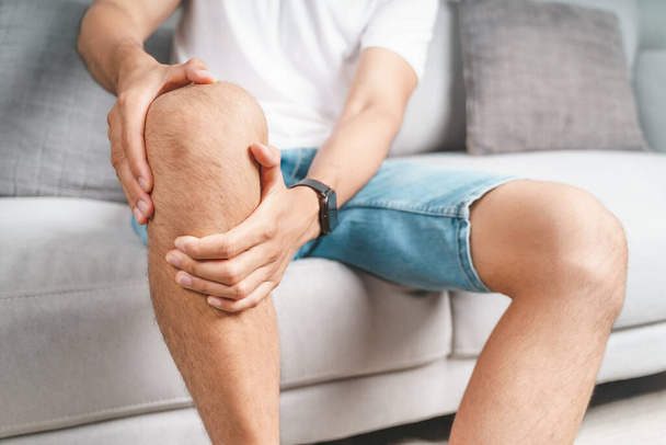 Мужчина, страдающий от боли в колене, сидит на диване и массирует больное колено.. - Фото, изображение