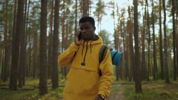 A sárga kabátos fekete férfi átsétál az erdőn és telefonon beszél egy lassított felvételű hátizsákkal.  - Felvétel, videó