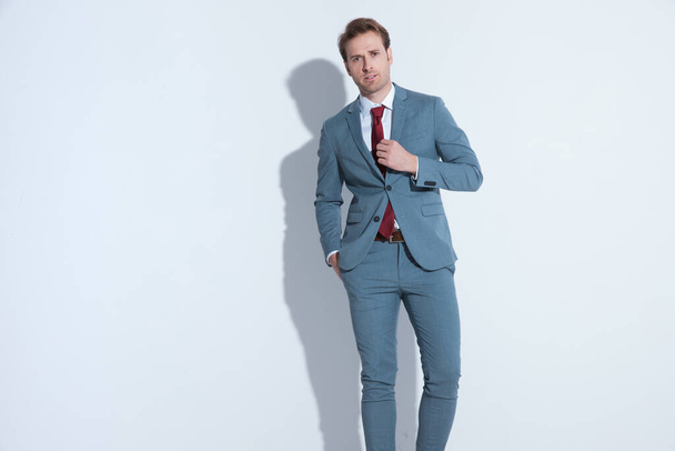 atractivo hombre de negocios sin afeitar en traje azul de la mano en el bolsillo y la fijación de la corbata mientras camina sobre fondo gris en el estudio - Foto, Imagen