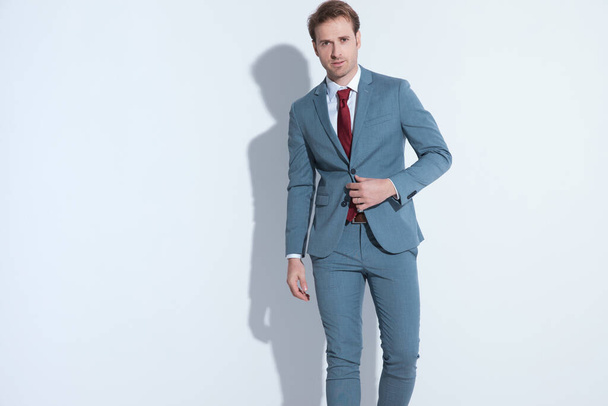 confident young guy buttoning and fixing blue elegant suit while walking and posing on gray background in studio - Foto, immagini