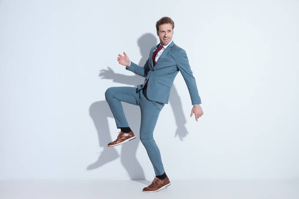 widok z boku szczęśliwy biznesmen w eleganckim niebieskim garniturze trzymając kolano do góry, śmiejąc się i bawiąc się podczas pozowania na szarym tle w studio - Zdjęcie, obraz