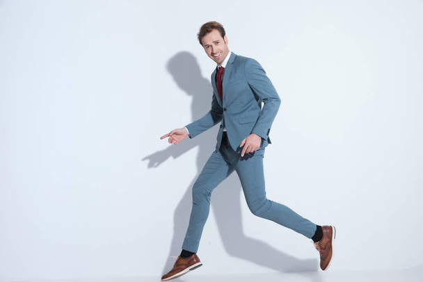 boldog fiatal férfi kék öltönyben ugrál a levegőben, ujjal mutogatva oldalra, mosolyogva és szórakozva szürke háttér a stúdióban - Fotó, kép