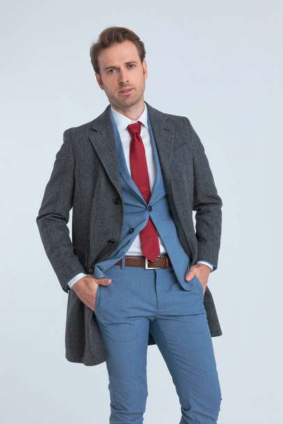 молодий модний бізнесмен стирчить своїми руками в кишенях, одягнений в синій костюм і пальто
 - Фото, зображення