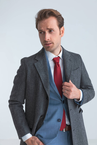 sexy zakenman rangschikken zijn jas, steken een hand in zak en kijken weg  - Foto, afbeelding