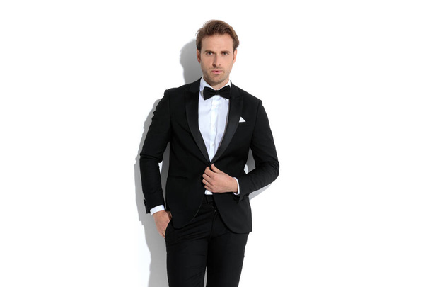 elegant businessman adjusting his tux and putting one hand in pocket against white background - Zdjęcie, obraz