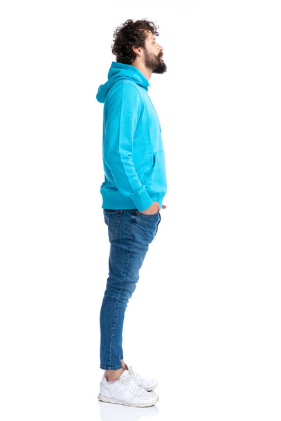full body foto van jong bebaarde man in blauw hoodie wachten in de rij terwijl poseren tegen witte achtergrond geïsoleerd in studio - Foto, afbeelding