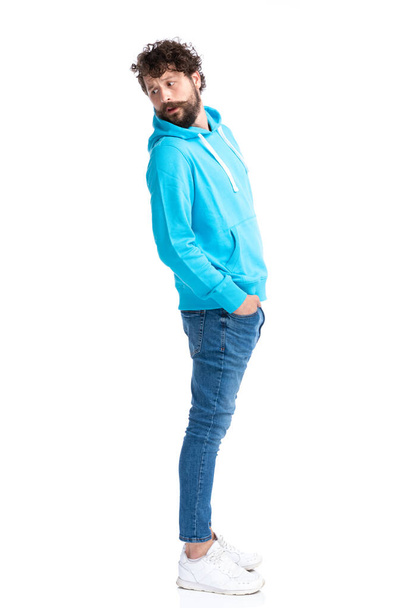 modello casual in felpa blu con cappuccio che tiene le mani in tasca e guarda curiosamente sopra la spalla mentre aspetta in fila su sfondo bianco in studio - Foto, immagini