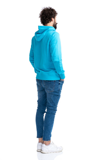 full body picture of unshaved young guy holding hands in pockets and waiting in line while standing against white background in studio - Foto, Imagem