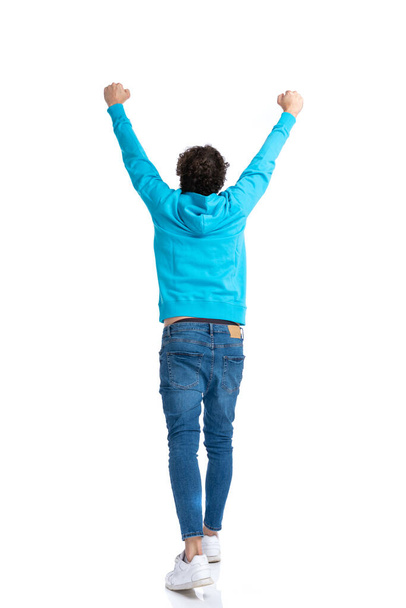 visão traseira do homem casual encaracolado com capuz azul segurando braços no ar e torcendo vitória, andando isolado no fundo branco no estúdio - Foto, Imagem