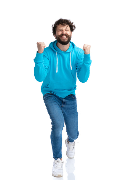 πλήρης εικόνα του σώματος του ενθουσιασμένοι γενειοφόρος μοντέλο μόδας σε μπλε hoodie κρατώντας τα χέρια και γιορτάζει τη νίκη, γέλιο και διασκέδαση σε λευκό φόντο στο στούντιο - Φωτογραφία, εικόνα