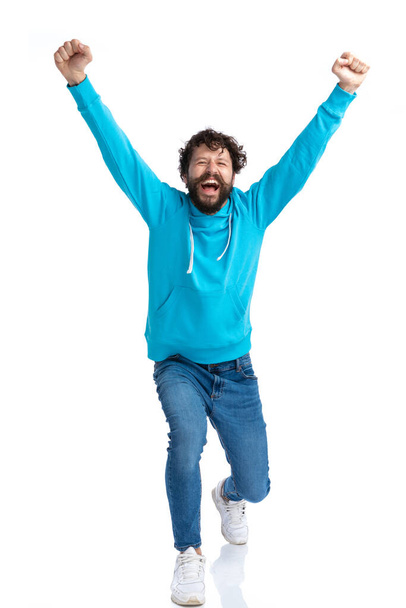 happy young man with long beard holding arms above head, laughing and celebrating victory while bending knee and posing against white background in studio - Photo, Image