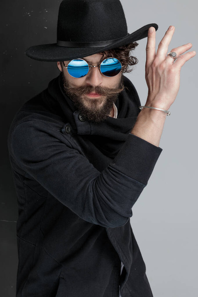 Dramatischer Typ mit langem Bart und Schnurrbart, der schwarzen Hut hochschiebt und cool vor schwarz-grauem Hintergrund im Studio posiert - Foto, Bild