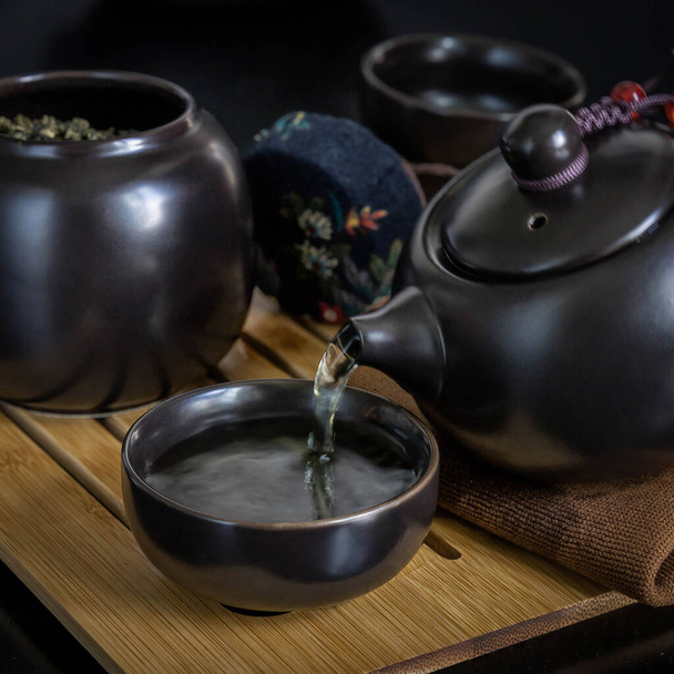 カップに緑茶を入れ、黒ティーポットと瓶を竹製のタブレットと黒の背景に醸造します。茶道 .  - 写真・画像