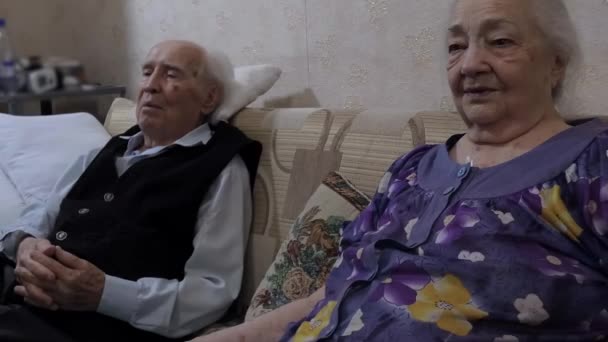 Een ouder stel, een man en een vrouw, zitten op de bank in hun huis. Ze kijken naar iets samen met rente. - Video