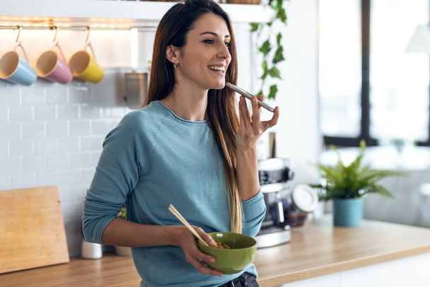 Shot of beautiful woman sending an audio messages with her smartphone while eating noodles with chopsticks in the kitchen at home. - Photo, Image