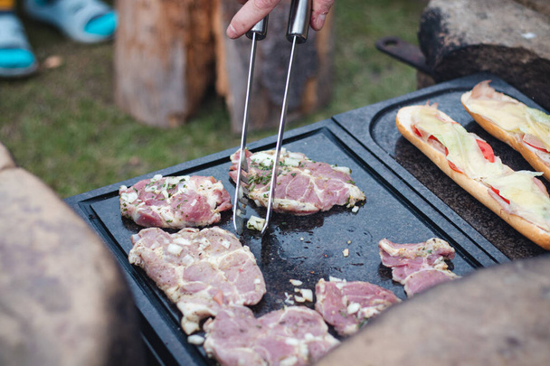 De mens bereidt een sappige varkenshals met basilicum en barbecue kruiden en een stokbrood met tomatenkaas en ham op een hete granieten plaat. Het barbecueseizoen is begonnen. Open haard. - Foto, afbeelding